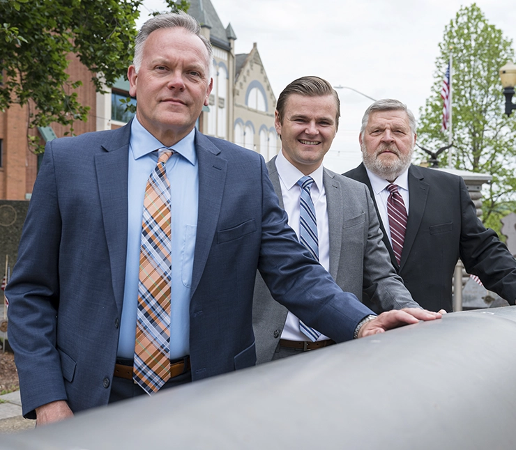 Three individuals in suits lean on a historical cannon in an outdoor setting, surrounded by greenery and buildings.