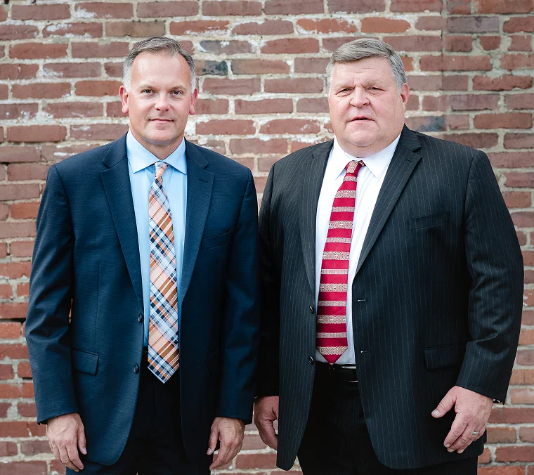 Three individuals in suits lean on a historical cannon in an outdoor setting, surrounded by greenery and buildings.