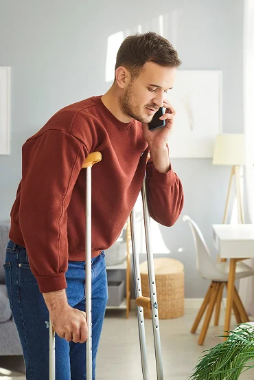 A person in a burgundy sweatshirt uses crutches while talking on the phone in a well-lit, modern living room.