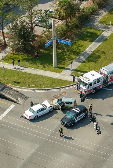Aerial view of a traffic accident involving two cars and emergency vehicles at an intersection, with bystanders observing nearby.