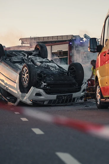 A flipped car on a highway with smoke rising, while emergency responders attend to the scene and a fire truck is visible in the background.