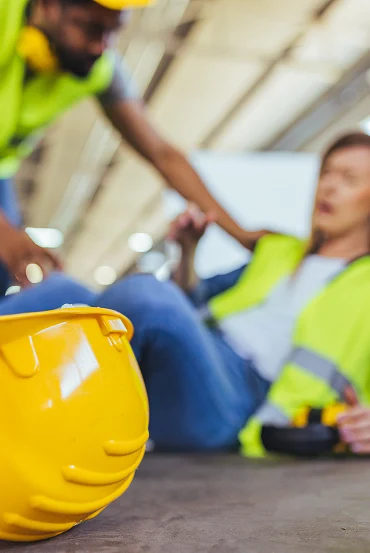 A yellow hard hat sits on the floor as a person reaches out to another who is laying on the ground in a workplace setting.