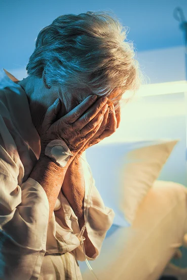 An elderly woman in a hospital gown covers her face with her hands, expressing distress against a softly lit background.